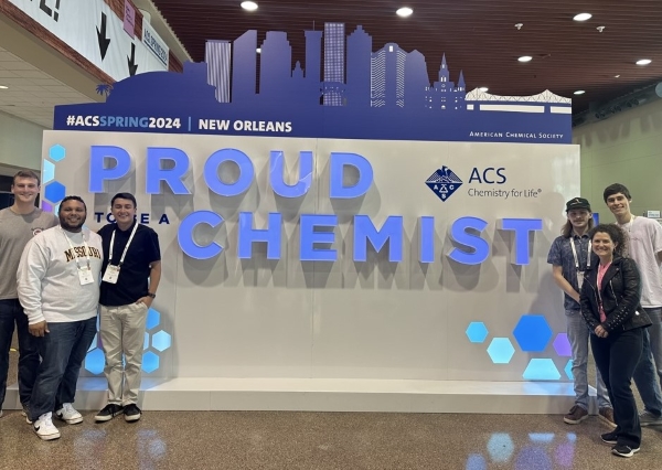 students and professor standing beside a "Proud Chemist" sign