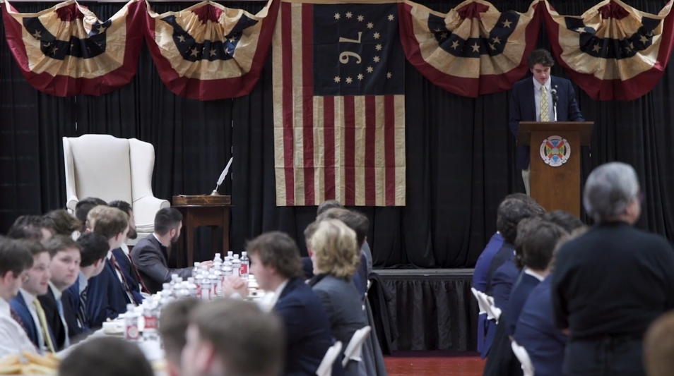 Screenshot of a video of a student speaking in front of his peer surrounded by American flags